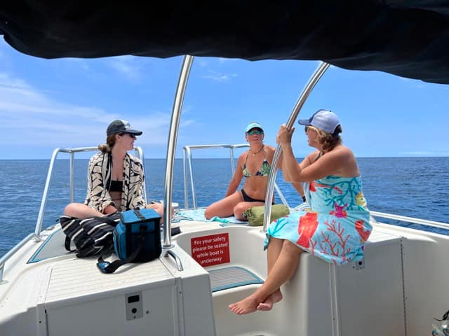 3 women sit on the bow of a boat in the sun talking