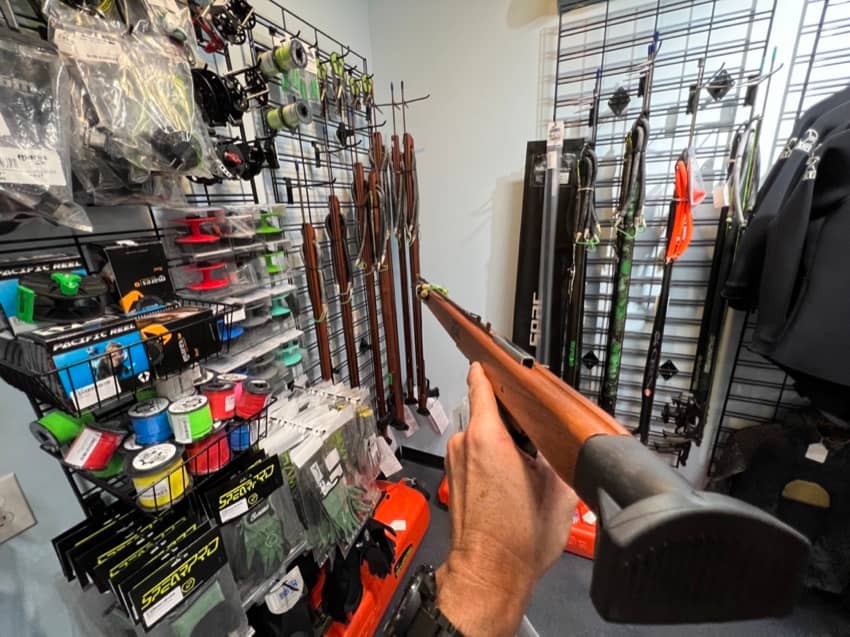 pointing a speargun at a wall display of other spearguns holding in hand first person