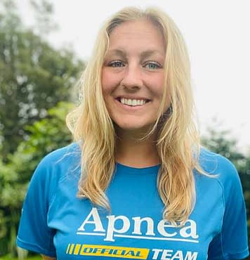 Blonde woman with long hair and blue eyes and nose ring wearing a turquoise shirt that says apnea freediving team smiling headshot