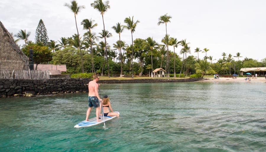 a man stands on a paddle board paddling while a woman sits in front. A Hawaiian heiau and beach are in the background and the water is calm and green