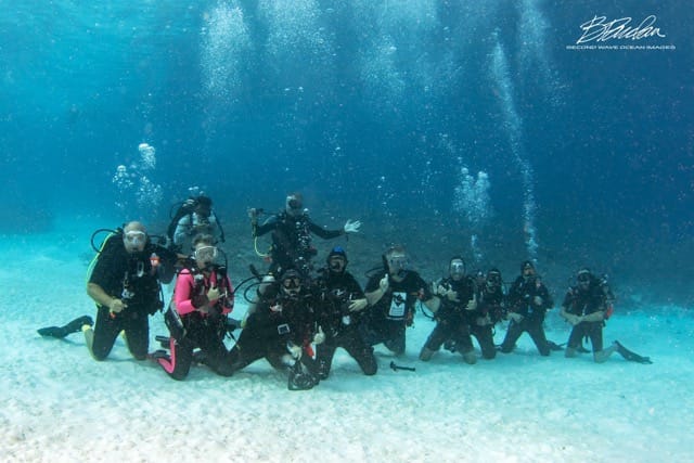 a group of 10 divers sitting on the bottom of the ocean in sand posing
