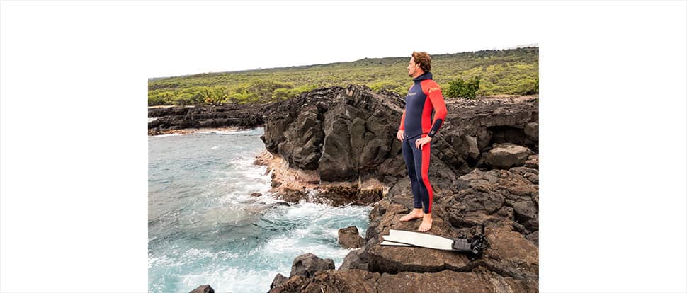 Byron Kay looks out over the ocean standing on kona lava coast in custom best dive wetsuit colored dark blue and orange