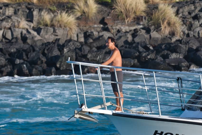 divemaster in swim trunks holds on to bow rail of boat