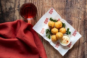 Salt cod and potato fritters on a white sheet of parchment paper served with a garnish of parsley, citrus aioli, and a beer.