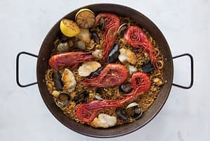 A dish made with bomba rice filled with seafood like monkfish, sepia, squid, shrimp, clams, and mussels. Garnished with saffron, lemon, and a salsa verde served in a large paella on a white surface.