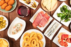 The food of Spain. An overhead photo of many different Spanish tapas, shot from above on a dark rustic texture. Jamon, cheese, wine, olives, croquettes, calamari rings, anchovies and more. Concept of Wine and Tapas pairing.