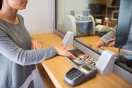 A woman at a bank, receiving money from the teller with a protective barrier between them