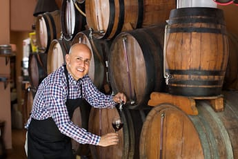 Smiling man in a winery with stacked barrels pouring a glass of red wine from a barrel with a spicket