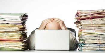 Man head behind a laptop with hands on bald head and a pile of paper files on either side