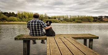 A man and a dog sitting on a pier looking out over a lake with a cloudy sky