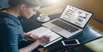 A woman sitting at a table in a coffee shop writing notes with coffee on her left, computer in front and phone on right