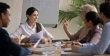 Group of people sitting around a table with a whiteboard and plant in the background