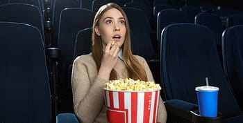 A woman seated watching a movie with a bucket of popcorn on her lap and drink in a blue cup