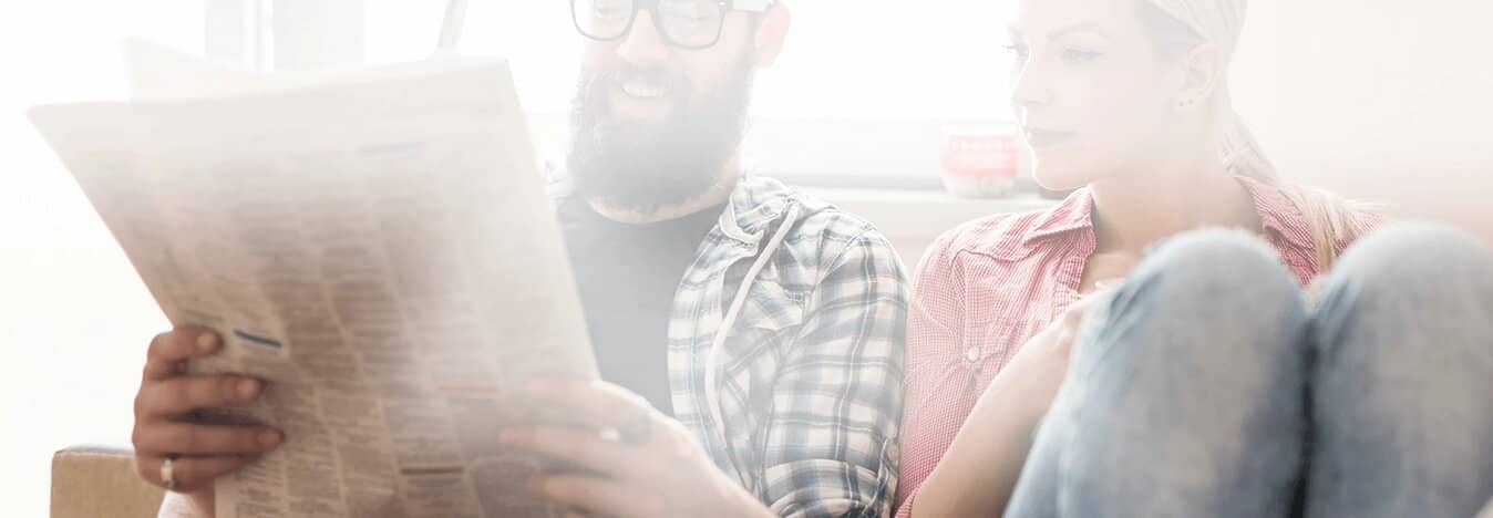 Two people sitting on a couch smiling and looking at a newspaper